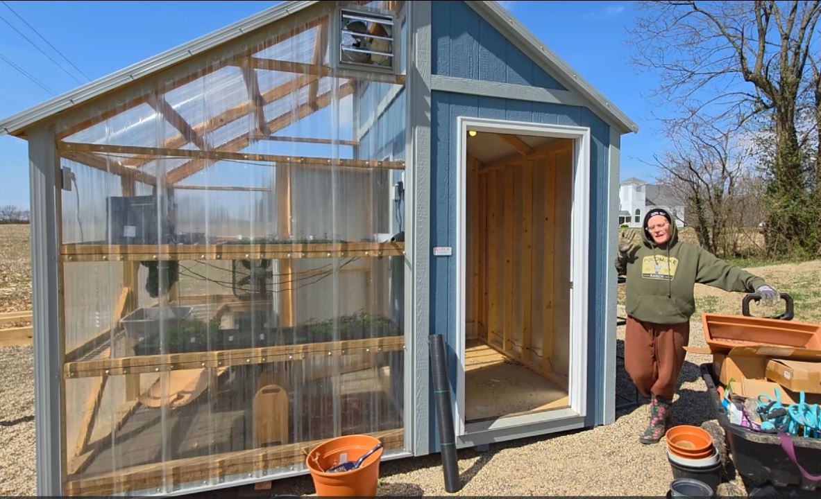 A woman stands next to a blue greenhouse and open utility shed during spring cleanup, smiling while holding a wheelbarrow full of gardening supplies.